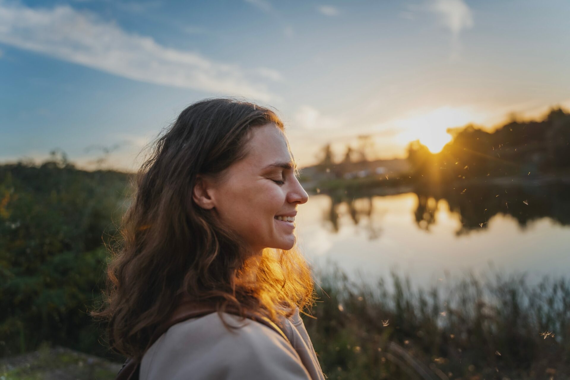 Une femme souriante profitant d'un coucher de soleil près d'un lac, reflétant la sérénité et la paix intérieure.