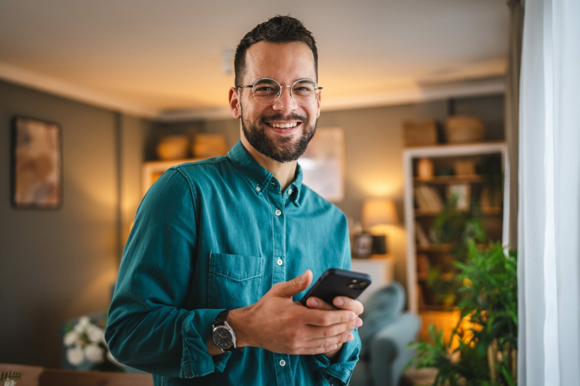 Un homme souriant tenant un smartphone dans un intérieur chaleureux et moderne.