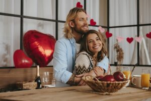 Couple souriant célébrant la Saint-Valentin à la maison avec décorations en forme de cœur, symbole d’un amour authentique et durable.